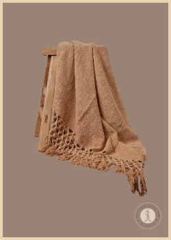 Tan woven cotton Layering Throw Blanket from Shoppe at The Avenue draped over a wood stool with crocheted ends and fringe.